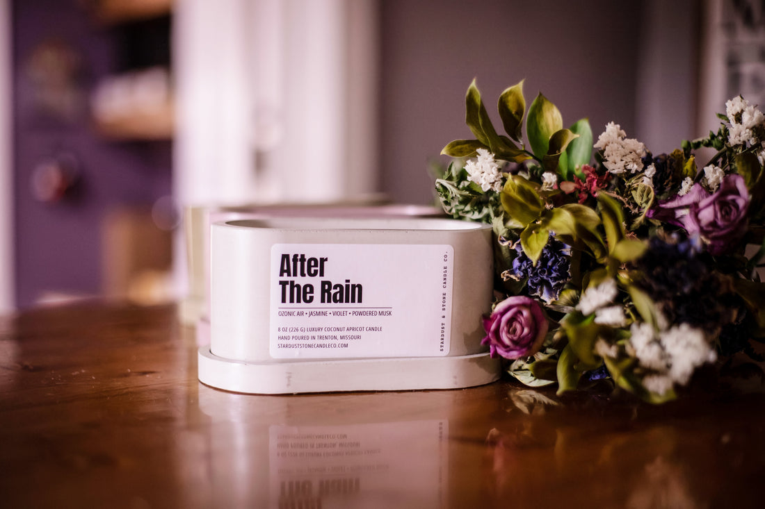 a grey concrete double wick candle in the scent After The Rain on a wooden countertop next to a dried floral bouquet and a blurred background.